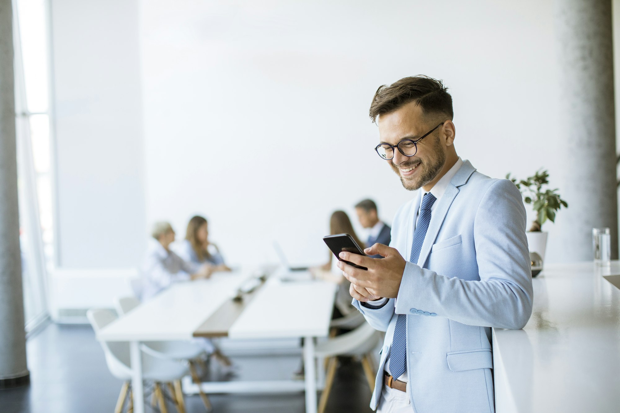 Happy young man using his mobile phone and smiling while his colleagues working in the background